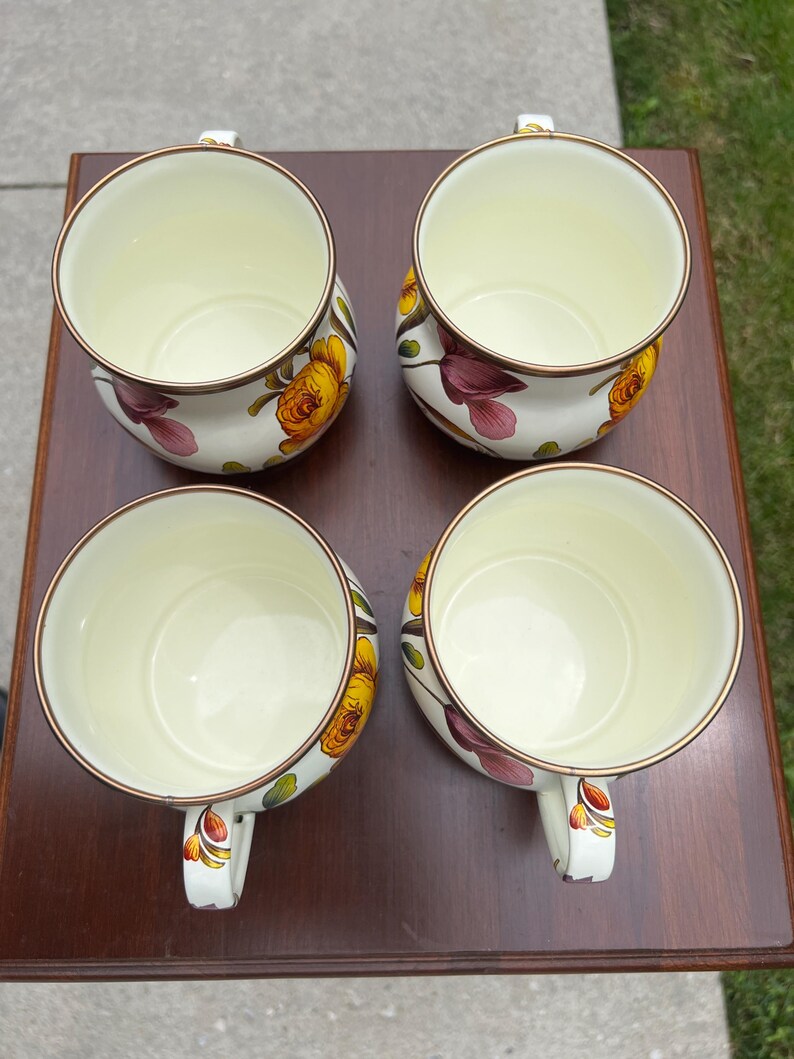 May include: A set of four cream-colored ceramic mugs with a floral pattern featuring yellow and purple flowers. Each mug has a rounded shape, a smooth interior, and a thin metallic-colored rim. The handles are decorated with small floral motifs.