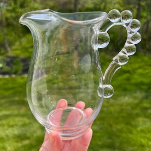 Peut inclure: Une carafe en verre transparent avec une poignée unique. La poignée est ornée d'une série d'éléments sphériques en verre transparent. La carafe a un bec évasé et un corps arrondi, parfait pour servir des boissons. La carafe est tenue devant un fond vert flou.
