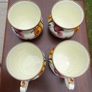 May include: A set of four cream-colored ceramic mugs with a floral pattern featuring yellow and purple flowers. Each mug has a rounded body, a single handle, and a thin metallic-toned rim. The interior of each mug is a smooth, solid cream color.