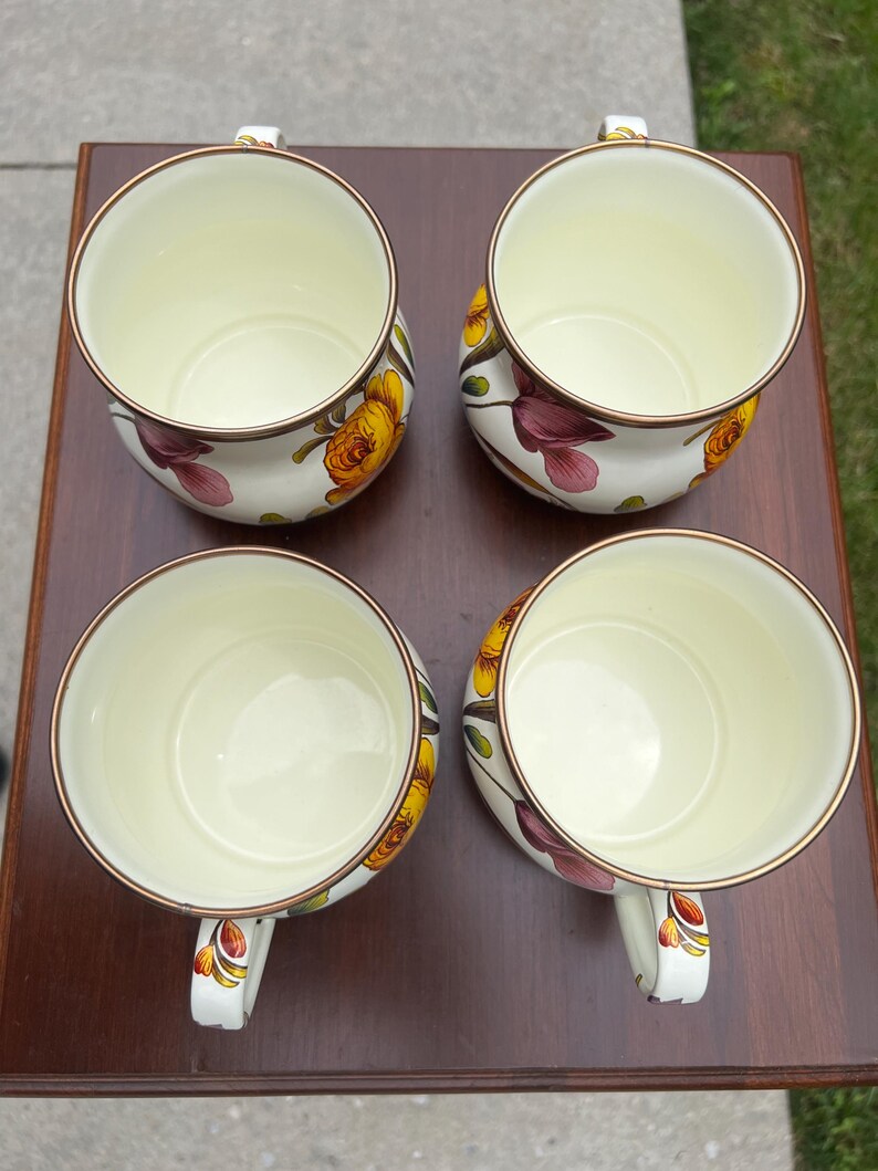 May include: A set of four cream-colored ceramic mugs with a floral pattern featuring yellow and purple flowers. Each mug has a rounded body, a single handle, and a thin metallic-toned rim. The interior of each mug is a smooth, solid cream color.
