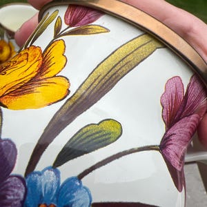 May include: A white enamel mug featuring a vibrant floral pattern with yellow, purple, and blue flowers and green leaves. The mug has a metallic bronze-colored rim and a smooth, glossy finish, showcasing detailed botanical illustrations on its surface.