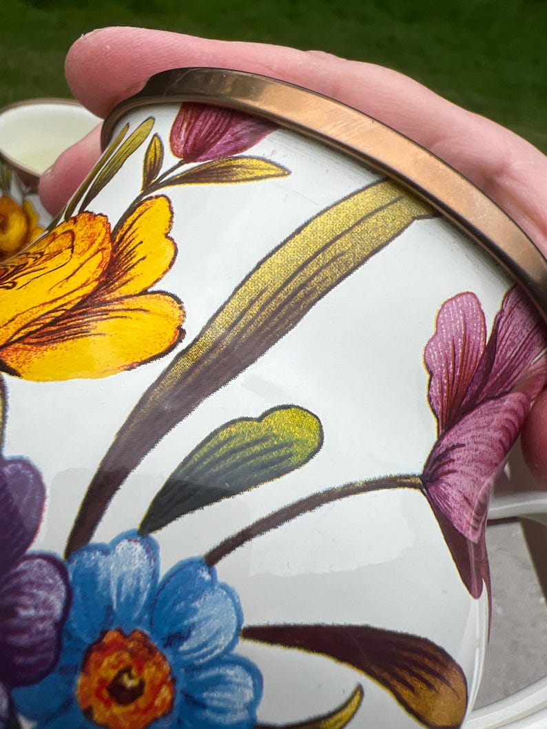 May include: A white enamel mug featuring a vibrant floral pattern with yellow, purple, and blue flowers and green leaves. The mug has a metallic bronze-colored rim and a smooth, glossy finish, showcasing detailed botanical illustrations on its surface.