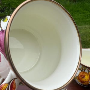 May include: A white ceramic mug with a smooth cream-colored interior and a metallic bronze-toned rim. The exterior features a floral pattern with pink and yellow petals and green leaves. The mug has a curved handle and a wide, circular opening.