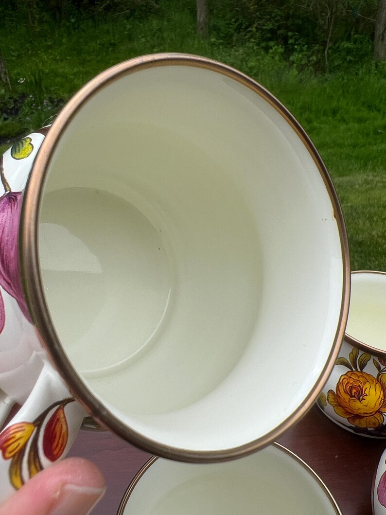 May include: A white ceramic mug with a smooth cream-colored interior and a metallic bronze-toned rim. The exterior features a floral pattern with pink and yellow petals and green leaves. The mug has a curved handle and a wide, circular opening.