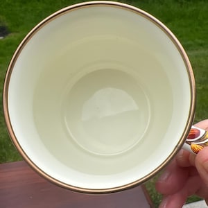 May include: A round ceramic mug with a smooth cream-colored interior and a thin metallic gold rim. The exterior handle features a decorative pattern with orange, brown, and yellow tones. The mug is viewed from directly above, showing the circular opening.
