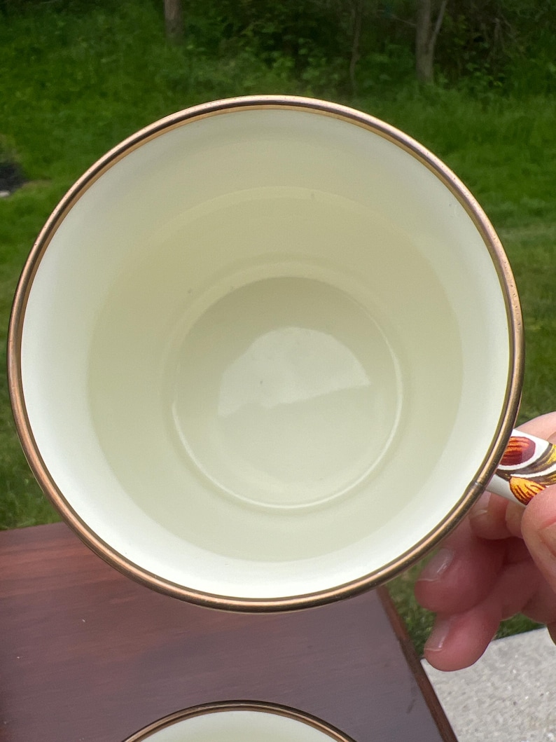 May include: A round ceramic mug with a smooth cream-colored interior and a thin metallic gold rim. The exterior handle features a decorative pattern with orange, brown, and yellow tones. The mug is viewed from directly above, showing the circular opening.