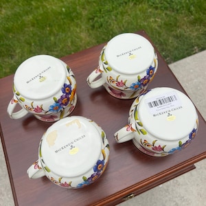 May include: Four white ceramic mugs with handles, each featuring a colorful floral pattern of blue, purple, and pink flowers. The undersides are marked with the MacKenzie-Childs logo, and one mug includes a barcode sticker with product text.