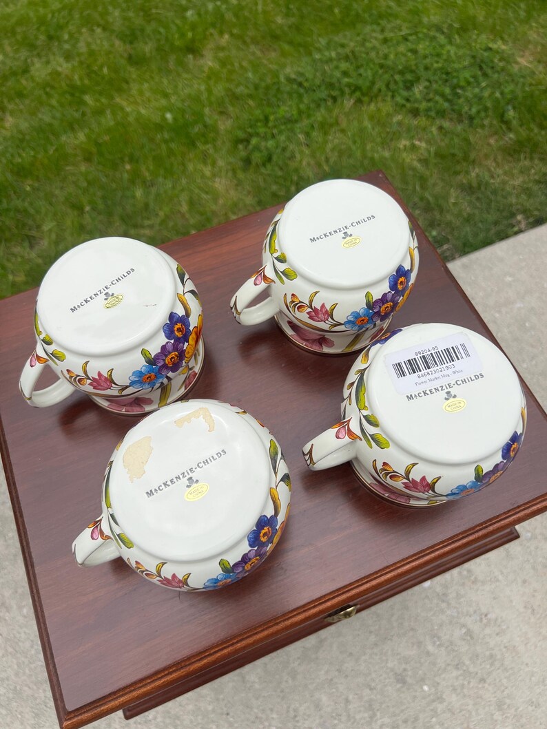 May include: Four white ceramic mugs with handles, each featuring a colorful floral pattern of blue, purple, and pink flowers. The undersides are marked with the MacKenzie-Childs logo, and one mug includes a barcode sticker with product text.