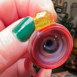 May include: A hand-blown glass art piece with a swirling vortex design. The piece features a vibrant color palette of red, purple, and blue, with a yellow top. The glass has a glossy finish and a unique, eye-catching shape. This piece is a decorative item.