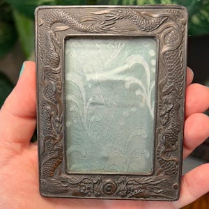 May include: Antique rectangular photo frame with a detailed dragon motif in a dark metallic finish. The frame's interior holds a light blue fabric with a subtle floral pattern, creating a vintage aesthetic. The frame is held in a hand.