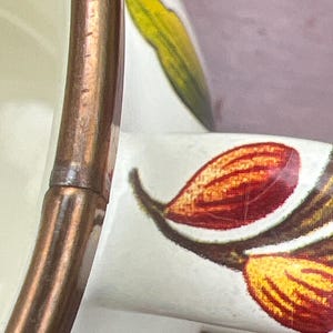May include: A white ceramic mug featuring a metallic copper-colored rim and a handle decorated with a botanical pattern of red, orange, and yellow almond-shaped elements alongside a green leaf, showing fine surface crazing on the glaze.
