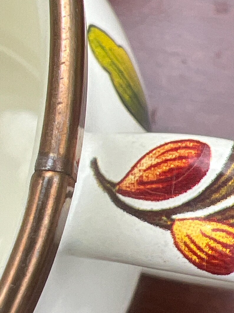 May include: A white ceramic mug featuring a metallic copper-colored rim and a handle decorated with a botanical pattern of red, orange, and yellow almond-shaped elements alongside a green leaf, showing fine surface crazing on the glaze.