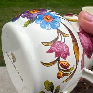 May include: A white ceramic mug featuring a colorful floral pattern with purple, blue, and pink flowers, green leaves, and gold-toned accents. The base of the mug displays the text MacKenzie-Childs and a small circular gold sticker label.