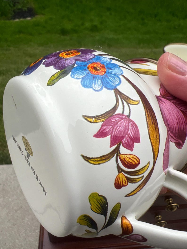 May include: A white ceramic mug featuring a colorful floral pattern with purple, blue, and pink flowers, green leaves, and gold-toned accents. The base of the mug displays the text MacKenzie-Childs and a small circular gold sticker label.