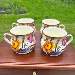 May include: A set of four white ceramic mugs featuring a colorful floral pattern with yellow, pink, and blue flowers, green leaves, and a metallic gold-toned rim. Each mug has a rounded body, a curved handle, and a smooth, glossy finish.