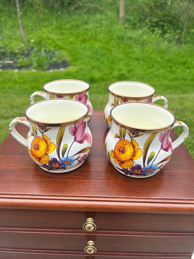 May include: A set of four white ceramic mugs featuring a colorful floral pattern with yellow, pink, and blue flowers, green leaves, and a metallic gold-toned rim. Each mug has a rounded body, a curved handle, and a smooth, glossy finish.