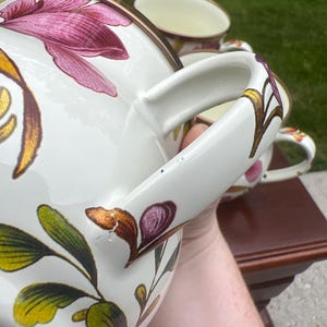 May include: A set of cream-colored ceramic mugs featuring a floral pattern with magenta, gold, and green leaves. Each mug has a curved handle and a metallic-toned rim. The design includes detailed botanical illustrations painted on the exterior surface.
