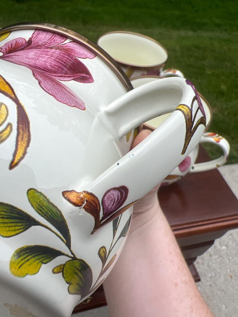 May include: A set of cream-colored ceramic mugs featuring a floral pattern with magenta, gold, and green leaves. Each mug has a curved handle and a metallic-toned rim. The design includes detailed botanical illustrations painted on the exterior surface.