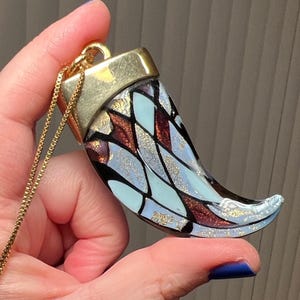 May include: A gold-toned necklace featuring a pendant shaped like a horn. The pendant has a gold cap and is decorated with a pattern of light blue, brown, and black shapes with gold flecks. The chain is a delicate gold color.