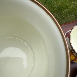 May include: A large, cream-colored bowl with a smooth interior and a metallic bronze-toned rim. Beside it, a smaller matching cup is visible, featuring a floral pattern with yellow and pink petals on its exterior surface.
