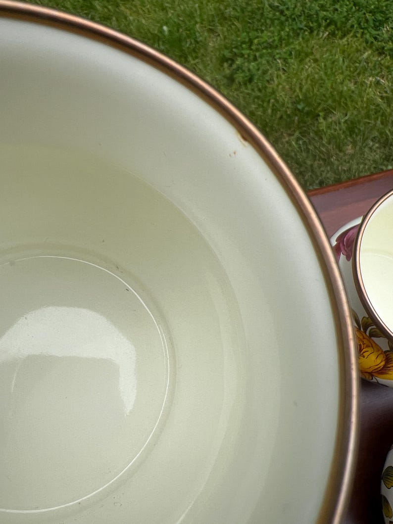 May include: A large, cream-colored bowl with a smooth interior and a metallic bronze-toned rim. Beside it, a smaller matching cup is visible, featuring a floral pattern with yellow and pink petals on its exterior surface.
