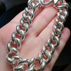 May include: A polished silver chain bracelet featuring thick, rounded interlocking links and a circular toggle clasp closure. The metal surface is highly reflective, showing smooth, curved edges throughout the entire length of the jewelry piece.