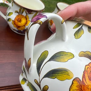 May include: A set of white ceramic mugs featuring a vibrant floral pattern with large yellow and orange roses, green leaves, and purple accents. Each mug has a curved handle and a metallic gold-toned rim, showcasing a detailed botanical design.