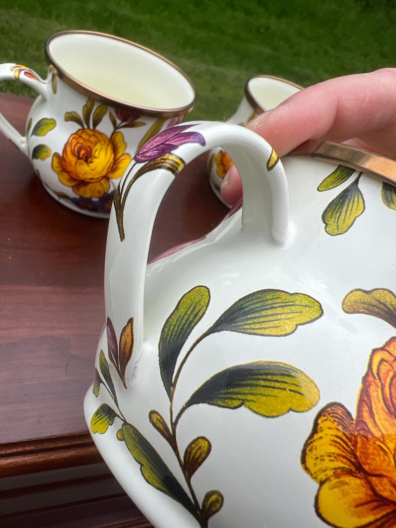 May include: A set of white ceramic mugs featuring a vibrant floral pattern with large yellow and orange roses, green leaves, and purple accents. Each mug has a curved handle and a metallic gold-toned rim, showcasing a detailed botanical design.