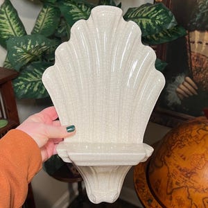 Puede incluir: Estante de pared de cerámica blanco roto con forma de concha. El estante tiene una parte superior festoneada y un pequeño estante rectangular. La superficie tiene un esmalte craquelado. El estante está sostenido por una base decorativa.