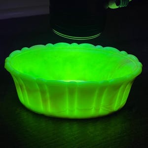 May include: A glowing green glass bowl with a scalloped rim and vertical ribbed sides. The bowl is illuminated, showcasing its vibrant color and design. The bowl is likely vintage or antique.