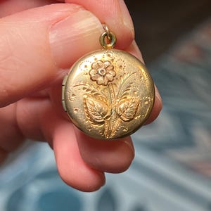 May include: A round gold-toned metal locket featuring an embossed floral design on the front. The centre of the locket displays a single flower with two leaves, surrounded by small engraved dots and decorative line patterns. A small metal loop is attached to the top.