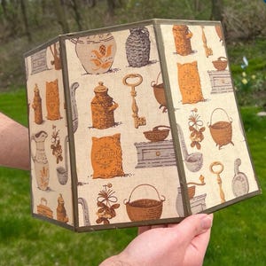 May include: A vintage lampshade with a beige background and a repeating pattern of kitchen-themed illustrations in brown and orange. The illustrations include keys, flour sacks, and jars. The lampshade has a rectangular shape.