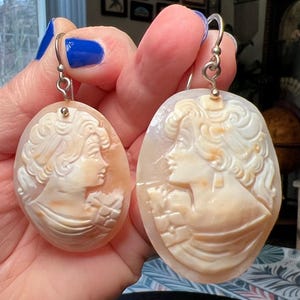 May include: A pair of oval cameo earrings. Each earring features a carved profile of a woman with flowing hair and classical attire. The earrings are a light peach colour with silver hooks. The cameo design is carved into the stone.