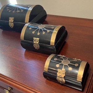 May include: Three black decorative boxes with brass accents. Each box has a hinged lid, a brass clasp, and a sunburst design on the top. The boxes are different sizes and sit on a dark wood surface.