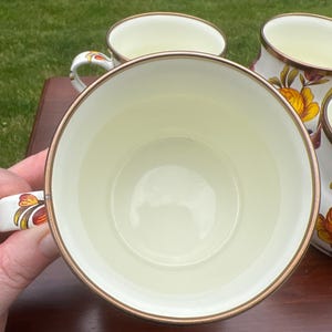May include: A set of cream-colored ceramic mugs featuring a floral pattern with orange and yellow petals and green leaves. Each mug has a metallic gold-toned rim and a matching floral design on the handle, providing a consistent look.
