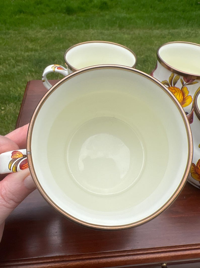 May include: A set of cream-colored ceramic mugs featuring a floral pattern with orange and yellow petals and green leaves. Each mug has a metallic gold-toned rim and a matching floral design on the handle, providing a consistent look.