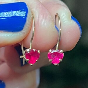 May include: A pair of gold-colored drop earrings, each featuring a heart-shaped, vibrant pink gemstone. The earrings are held up, showcasing their design and color against a blurred background.