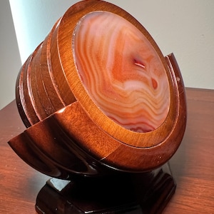 May include: A decorative coaster set featuring a polished agate stone centre with warm orange and white banding, set within a dark wooden frame. The coaster set is displayed on a dark wooden base, showcasing a unique design.