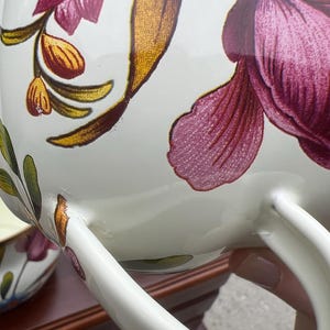 May include: A white ceramic teapot or pitcher featuring a floral pattern with large magenta petals, golden-brown leaves, and green stems. The vessel has a curved handle and a smooth, glossy finish, with a matching smaller piece visible in the background.