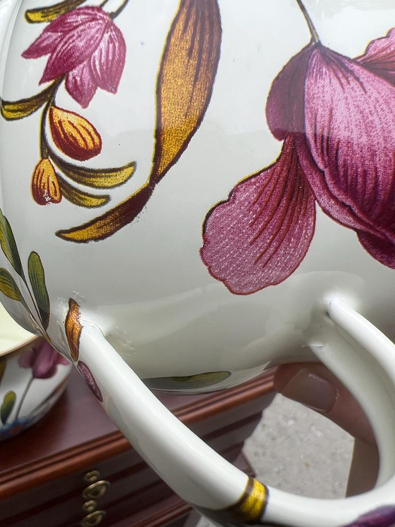 May include: A white ceramic teapot or pitcher featuring a floral pattern with large magenta petals, golden-brown leaves, and green stems. The vessel has a curved handle and a smooth, glossy finish, with a matching smaller piece visible in the background.