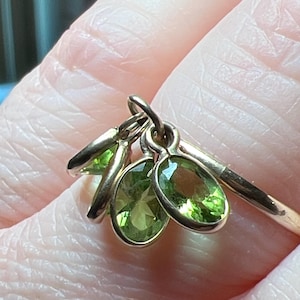 May include: A gold ring featuring three dangling oval-shaped charms, each set with a faceted, light green peridot gemstone. The charms are suspended from a thin, polished gold band, creating a layered look with the vibrant green stones.