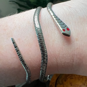 Op de afbeelding: Een zilverkleurige metalen slangenarmband met een gestructureerd schubpatroon langs het lichaam en twee kleine, felrode cirkelvormige accenten die de ogen op de kop voorstellen. Het ontwerp wikkelt zich in een spiraalvorm om de pols.