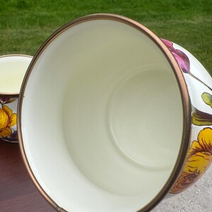 May include: A white enamel mug with a metallic bronze rim, featuring a vibrant floral pattern with yellow, orange, and purple flowers on the exterior. The interior of the mug is a smooth, solid cream color, and the handle is also decorated.