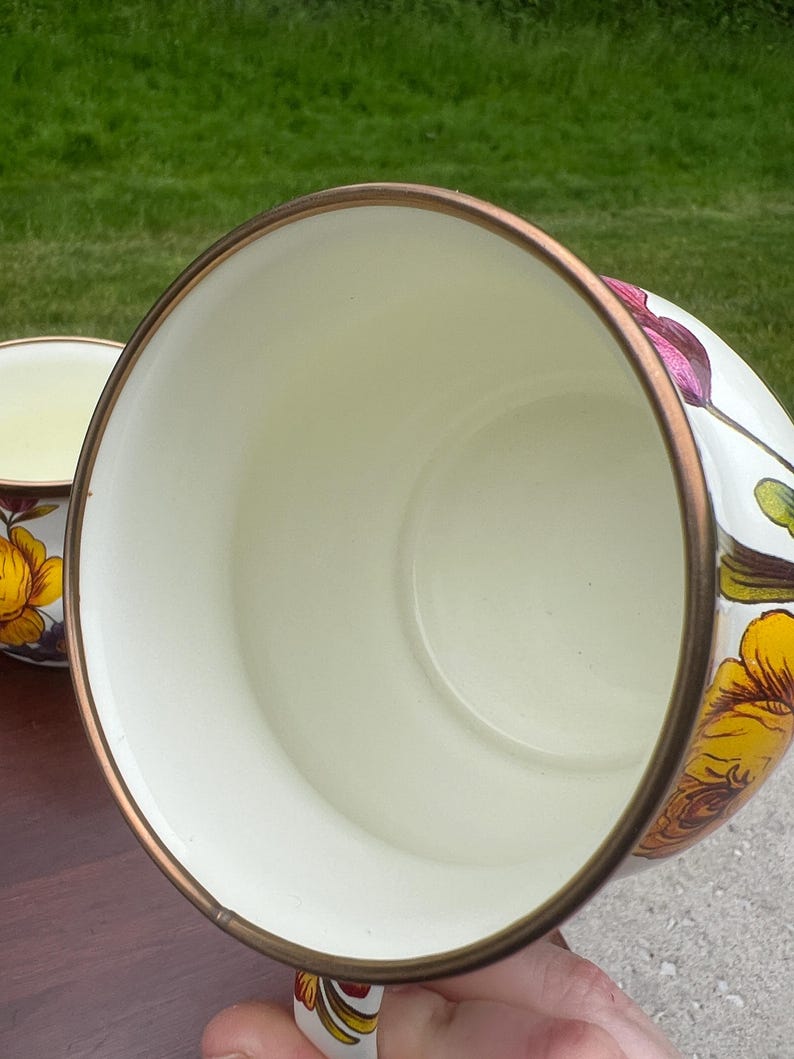 May include: A white enamel mug with a metallic bronze rim, featuring a vibrant floral pattern with yellow, orange, and purple flowers on the exterior. The interior of the mug is a smooth, solid cream color, and the handle is also decorated.
