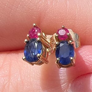 Könnte beinhalten: Ein Paar Ohrstecker in Gold, wobei jeder Ohrstecker einen oval geschliffenen blauen Saphir unter einem kleineren, rund geschliffenen roten Rubin aufweist. Die Steine sind in Krappenfassungen gefasst und bilden eine vertikale Anordnung.