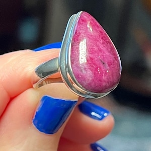 May include: A silver ring featuring a large, teardrop-shaped, pink gemstone. The gemstone has a marbled appearance with darker pink veins. The ring's band is a simple, polished silver.