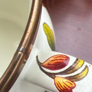 May include: A white ceramic mug featuring a metallic copper-colored rim and a handle decorated with a botanical pattern of red, yellow, and green leaves. The interior of the mug is cream-colored, and the exterior surface displays a smooth finish.
