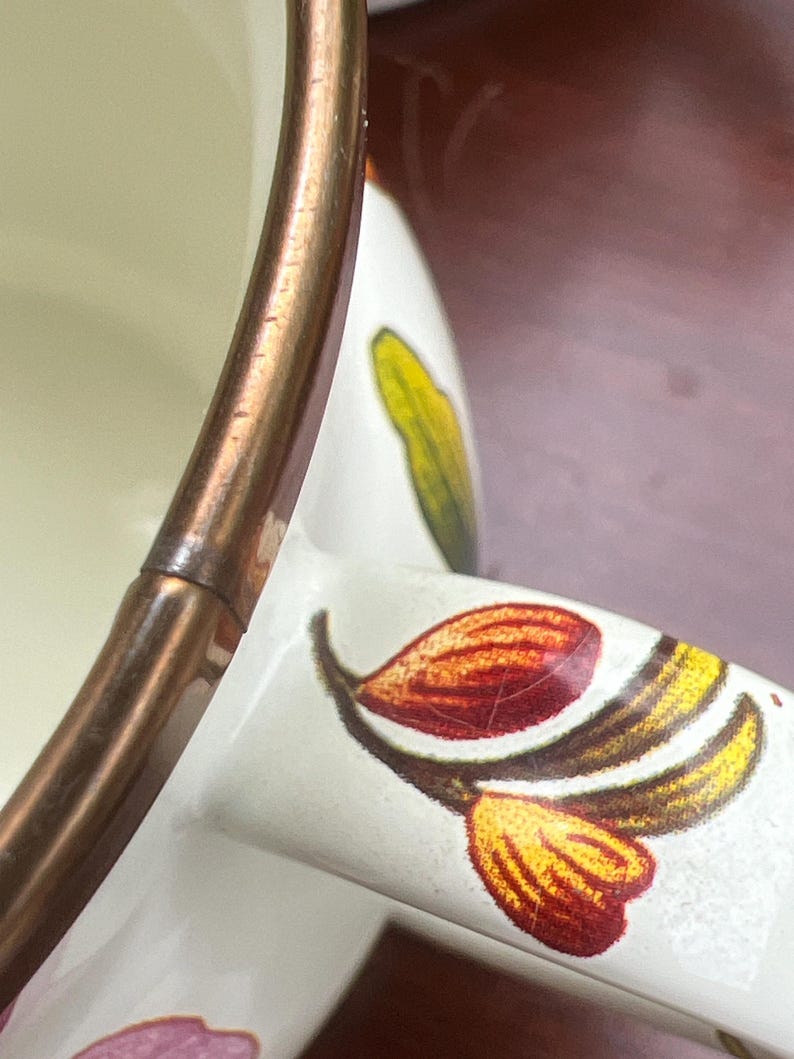 May include: A white ceramic mug featuring a metallic copper-colored rim and a handle decorated with a botanical pattern of red, yellow, and green leaves. The interior of the mug is cream-colored, and the exterior surface displays a smooth finish.