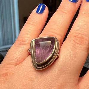 May include: A silver ring featuring a large, triangular amethyst gemstone. The amethyst has a deep purple hue and is set in a silver bezel. The ring has a wide band and a simple, elegant design, suitable for everyday wear.
