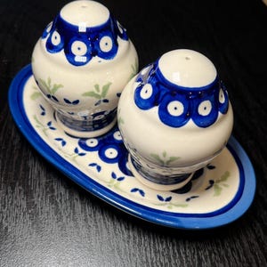 May include: A pair of cream-colored ceramic salt and pepper shakers with a rounded shape, featuring a blue and white circular pattern around the top and small green leaf accents, resting on a matching oval-shaped ceramic tray with a blue rim.